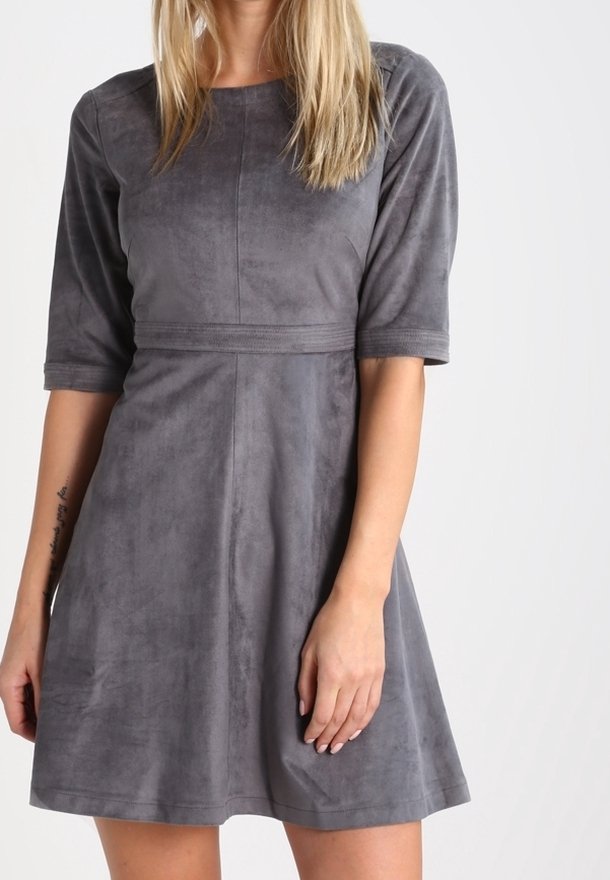 Femme portant une robe en daim grise à manches courtes, longueur genou, cintrée à la taille, sur un fond blanc uni.