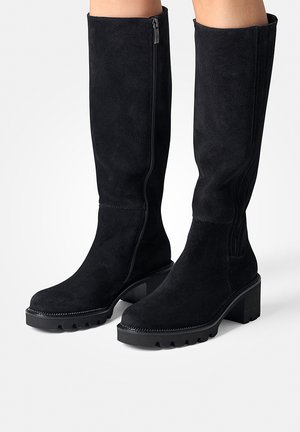 Las botas negras de ante hasta la rodilla cuentan con un cierre lateral, un tacón grueso negro y una suela de goma texturizada con dibujo. Resaltadas por sutiles detalles de costura.