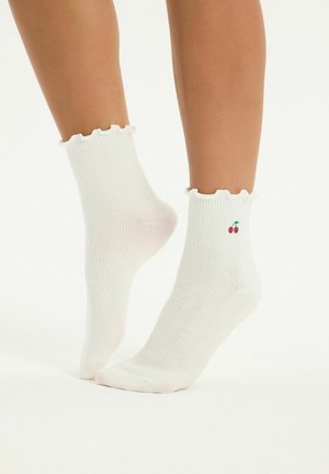 Stopy w białych prążkowanych skarpetach do kostki z falbankowymi ściągaczami i małym haftem wiśni po zewnętrznej stronie każdej skarpety.