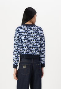 Knitted sweater with a blue heart pattern, cropped design, ribbed cuffs and hem, worn over dark denim pants with a logo patch on the back.