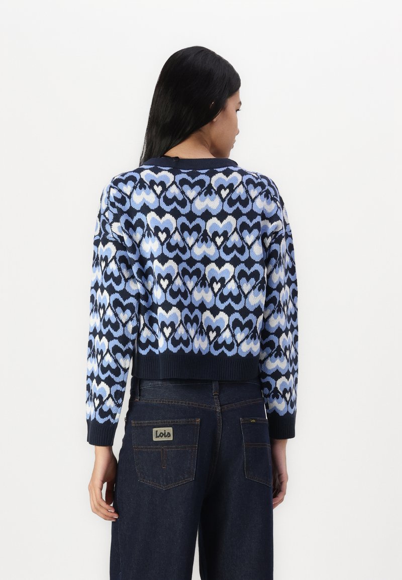 Gebreide trui met een blauw hartpatroon, cropped ontwerp, geribde manchetten en zoom, gedragen over donkere spijkerbroeken met een logo-opdruk op de achterkant.