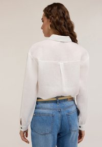 Camicia bianca leggera con bottoni, maniche lunghe e colletto, indossata con jeans in denim blu. La parte posteriore presenta uno yoke e una piccola piega.