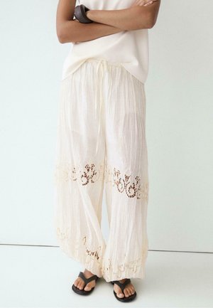 Donna che indossa pantaloni bianchi larghi con inserti in pizzo vicino a ginocchia e caviglie, abbinati a sandali neri e a un top bianco senza maniche.
