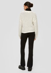 Weißer Strickpullover mit strukturiertem Muster, kombiniert mit schwarzen, gerippten, ausgestellt geschnittenen Hosen. Das Outfit hat eine lockere Passform und dezente Details.
