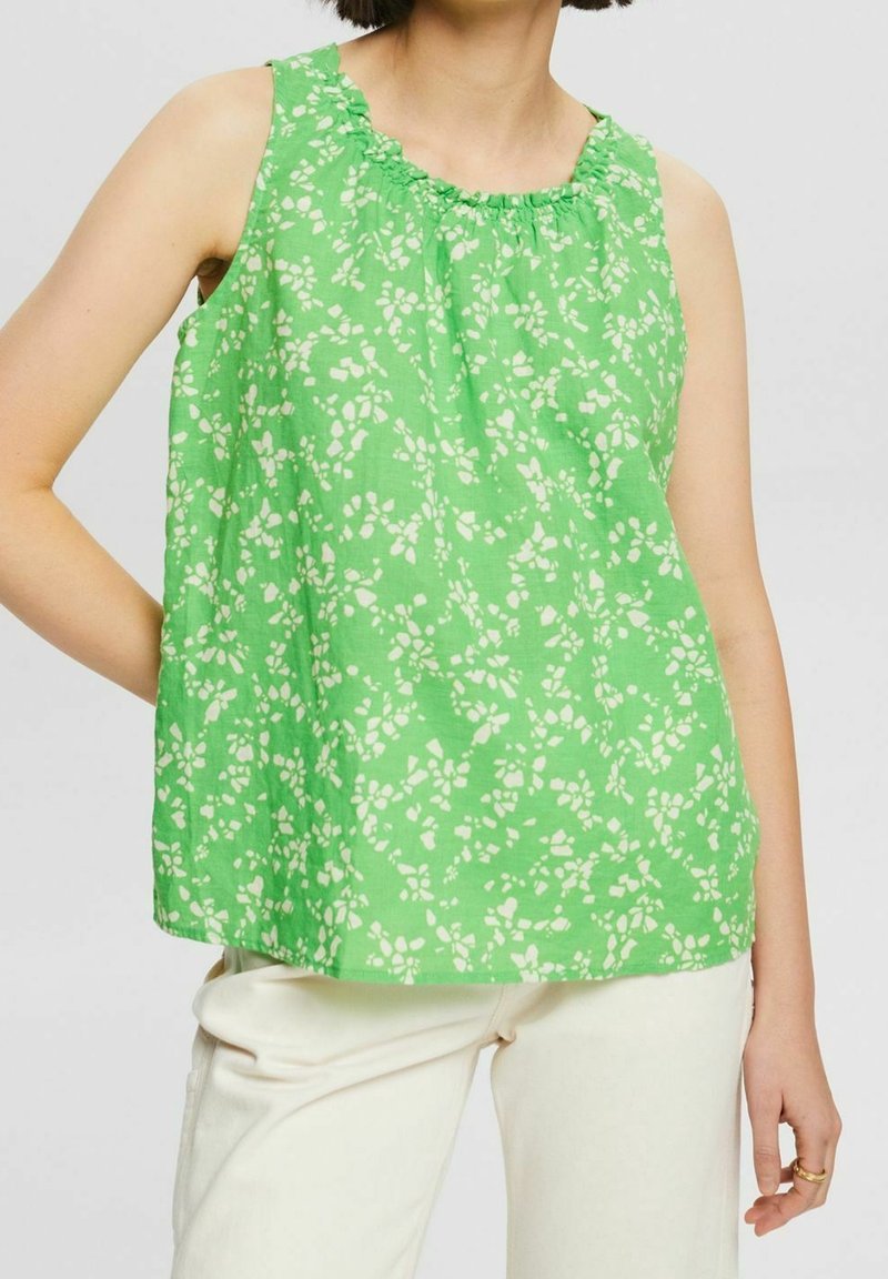 Femme portant un haut vert sans manches à motif floral blanc et un pantalon écru, debout avec une main sur la hanche devant un fond uni.