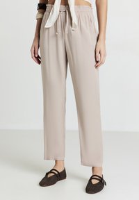 Donna che indossa pantaloni beige larghi con vita regolabile e scarpe basse intrecciate scure con doppie cinghie e fibbie.