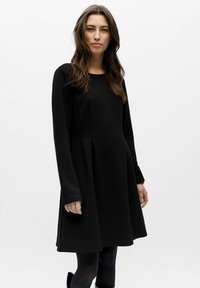 Robe noire à manches longues avec un col arrondi, une silhouette trapèze et des détails plissés. Confectionnée dans un tissu doux et texturé.