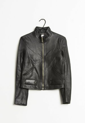 Czarna skórzana kurtka z frontowym zamkiem błyskawicznym, wysokim kołnierzem, dwoma bocznymi kieszeniami oraz zapinanymi na guziki paskami na dole, wisząca na czarnym wieszaku.