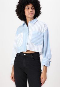 Klippt skjorta med knappar i ljusblått med vita ränder, med två framfickor och uppvikta ärmar, i kombination med svarta högmidjade jeans.