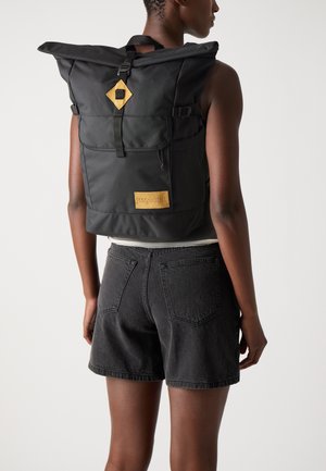 Sac à dos roll-top en tissu noir avec des bretelles réglables, poche avant et patch en cuir marron. Texture lisse avec un design minimaliste.