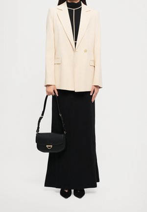 Kobieta ubrana w kremową marynarkę na czarnej długiej sukience, trzymająca czarną skórzaną torebkę z złotym zapięciem, stojąca na jednolitym tle.