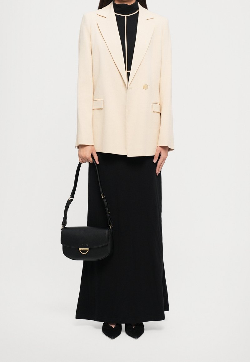 Femme portant un blazer crème sur une longue robe noire, tenant un sac à main en cuir noir avec fermoir doré, debout devant un fond uni.