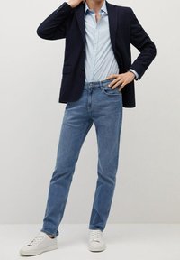 Granatowa marynarka na jasnoniebieskiej koszuli z guzikami, zestawiona z jasnoniebieskimi dżinsami i białymi trampkami. Gładkie tekstury, dopasowany krój i casualowy styl.