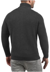Kullsvart ribbestrikket genser med høy krage, med en tykk tekstur og tettsittende ermer. Enkel design, ingen synlige dekorasjoner.
