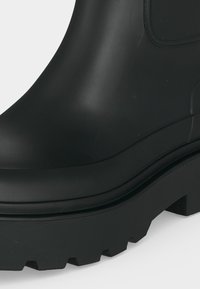 Gros plan d'une botte en caoutchouc noire avec une semelle épaisse et un profond motif de la bande de roulement sur fond blanc.
