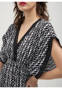 Blouse à motifs noir et blanc avec un décolleté en V profond, des manches courtes et une taille froncée. Présente des finitions en dentelle le long de l'encolure et des manches.