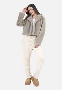 Kobieta stoi ubrana w beżową kurtkę z sztucznego futra, białą krótką bluzkę, kremowe spodnie z troczkami i beżowe buty slip-on na białym tle.