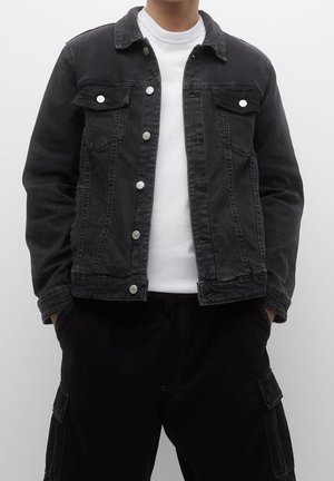 Personne portant une veste en jean noire sur un t-shirt blanc col rond avec un pantalon cargo noir, mains dans les poches devant un fond uni.