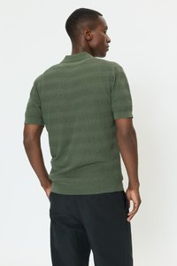 Polokošile s krátkým rukávem v olivově zelené barvě s texturovanými pruhy, žebrovaným límečkem a manžetami. Zobrazená zezadu, ve spojení s tmavými kalhotami.