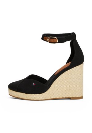 Sandale compensée noire avec bride à la cheville et boucle dorée, semelle plateforme beige tissée et petit logo rouge, blanc et bleu sur la zone des orteils.