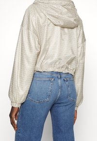 Persona che indossa una giacca con cappuccio beige con un motivo ripetuto "MK" e jeans in denim blu a vita alta, vista da dietro.