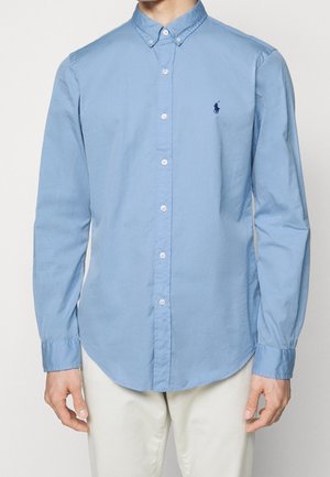 Chemise bleu clair à manches longues boutonnée avec col boutonné portée par une personne, associée à un pantalon beige clair, affichant le logo Polo Ralph Lauren.