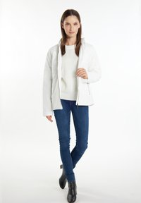 Hvid pufferjakke med høj krave og lynlås, lagdelt over en cremefarvet sweater. Båret med blå skinny jeans og sorte ankelstøvler.