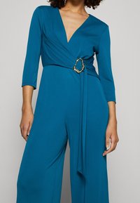 Combinaison teal avec un décolleté en V profond et des manches trois-quarts, dotée d'une boucle d'accent en or à la taille et d'une coupe évasée. Texture lisse.