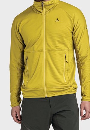Mann trägt eine gelbe Jacke mit Reißverschluss und zwei Reißverschlusstaschen sowie schwarze Hose, steht vor neutralem Hintergrund.