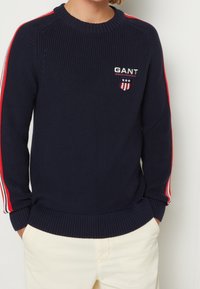 Navy gestrickter Pullover mit geripptem Ausschnitt und Bündchen, der rote und weiße gestreifte Akzente an den Ärmeln und ein gesticktes GANT-Logo aufweist.