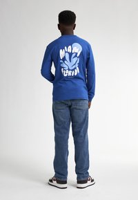 Langärmliges blaues Hemd mit weißem Aufdruck "MIAMI FLORIDA" und Spritzdesign auf dem Rücken, kombiniert mit blauen Jeans und schwarzen Sneakers.