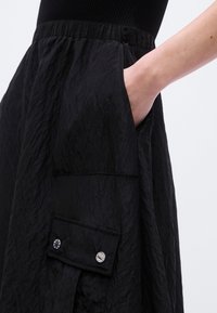 Χέρι που ακουμπάει μέσα στην πλαϊνή τσέπη μιας μαύρης υφασμάτινης φούστας με ρυτιδωμένη υφή και μια τσέπη με καπάκι και κουμπί από κάτω.