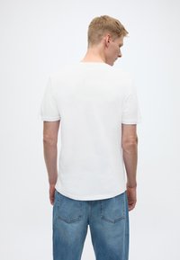 Bijela majica kratkih rukava, glatke teksture, zaobljenog izreza, s jednostavnim dizajnom, nošena uz svijetloplave široke traperice.