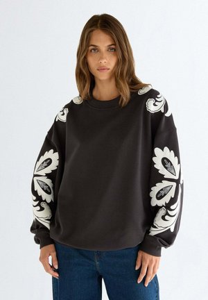 Frau trägt einen schwarzen oversized Sweatshirt mit großen weißen Blumenmustern auf den Ärmeln, kombiniert mit blauen Jeans, stehend vor einem weißen Hintergrund.