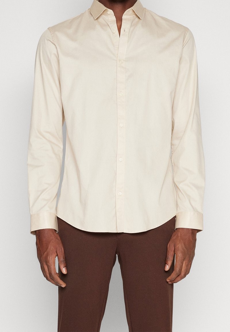 Beige long-sleeve shirt with a button-down collar, made of smooth fabric. Features buttons along the front and cuffed sleeves.