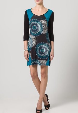 Robe à manches courtes avec une base noire, des accents teal et des motifs circulaires en blanc et turquoise. Fabriquée dans un tissu extensible, longueur genoux.