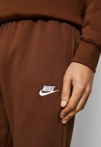 Detailní záběr ruky osoby odpočívající vedle hnědých tepláků Nike s bílým vyšitým logem na stehně.