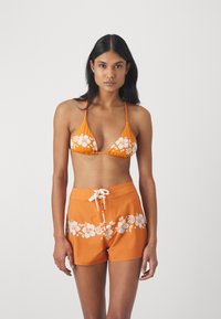 Haut de bikini orange avec un motif floral blanc, associé à un short de bain orange portant le même design. Bretelles réglables et lien à l'avant.