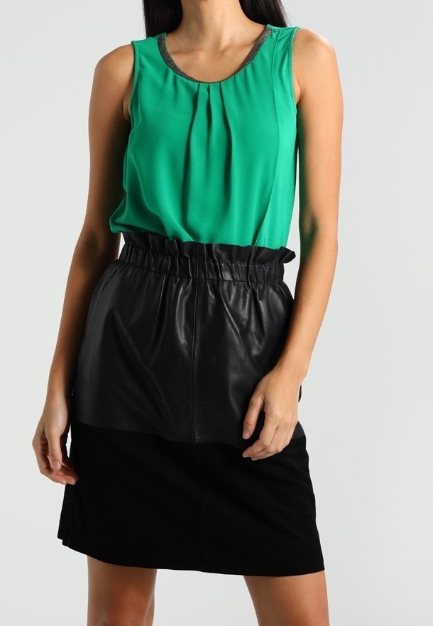 Top verde senza maniche con pieghe e rifiniture metalliche, abbinato a una gonna in pelle e camoscio nera con cintura elastica.