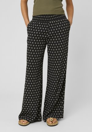 Personne portant un pantalon noir à jambes larges avec un motif losange blanc, un top vert sans manches, les mains dans les poches et des sandales beiges cloutées.