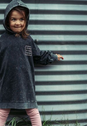 Dunkler, strukturierten Hoodie mit großer Kapuze und Fronttasche. Mit einem Logo-Patch in Hellpink. Kombiniert mit hellpinkfarbigen Leggings vor einer gewellten Oberfläche.