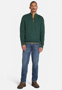 Grüner, gerippter Strickpullover mit einem Reißverschluss bis zum Kragen, kombiniert mit blauen Jeans und braunen Schnürstiefeln. Das Design zeichnet sich durch eine strukturierte Oberfläche aus.