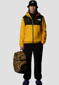 Gelbe und schwarze Jacke mit Reißverschluss, schwarze Hose, lila Hausschuhe und ein gemusterter Rucksack mit gelben und schwarzen Akzenten.