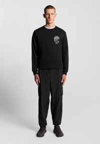 Felpa nera con design grafico bianco che presenta un leone e "1874" sul lato sinistro del petto, abbinata a pantaloni cargo neri e sneaker scure.