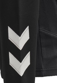 Svart atletisk topp med strukturerad tyg på ena sidan och slät material på den andra, med två vita chevronmönster.