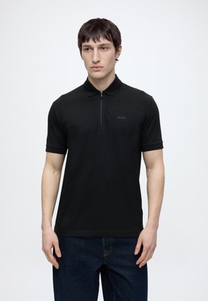 Fiatal férfi fekete rövid ujjú galléros pólót visel elöl cipzárral és sötétkék farmert, egyszínű fehér háttér előtt.