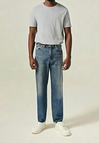 Homme portant une chemise à manches courtes gris clair, un jean bleu avec des détails délavés, et des baskets blanches sans lacets, debout devant un fond uni.