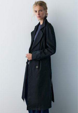 Vaaleahiuksinen nainen, jolla on pitkä musta trenssitakki tumman paidan päällä, seisoo yksinkertaisen vaaleanharmaan taustan edessä.