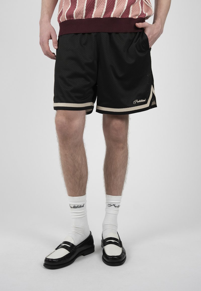 Personne portant un short en mesh noir avec un bord blanc, des chaussettes blanches avec un texte noir et des mocassins noir et blanc, debout devant un fond uni.