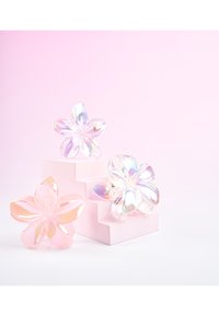 Trois pinces à cheveux en forme de fleur irisées, aux teintes roses et transparentes, exposées sur et à côté d'un petit piédestal à marches de couleur rose pâle.